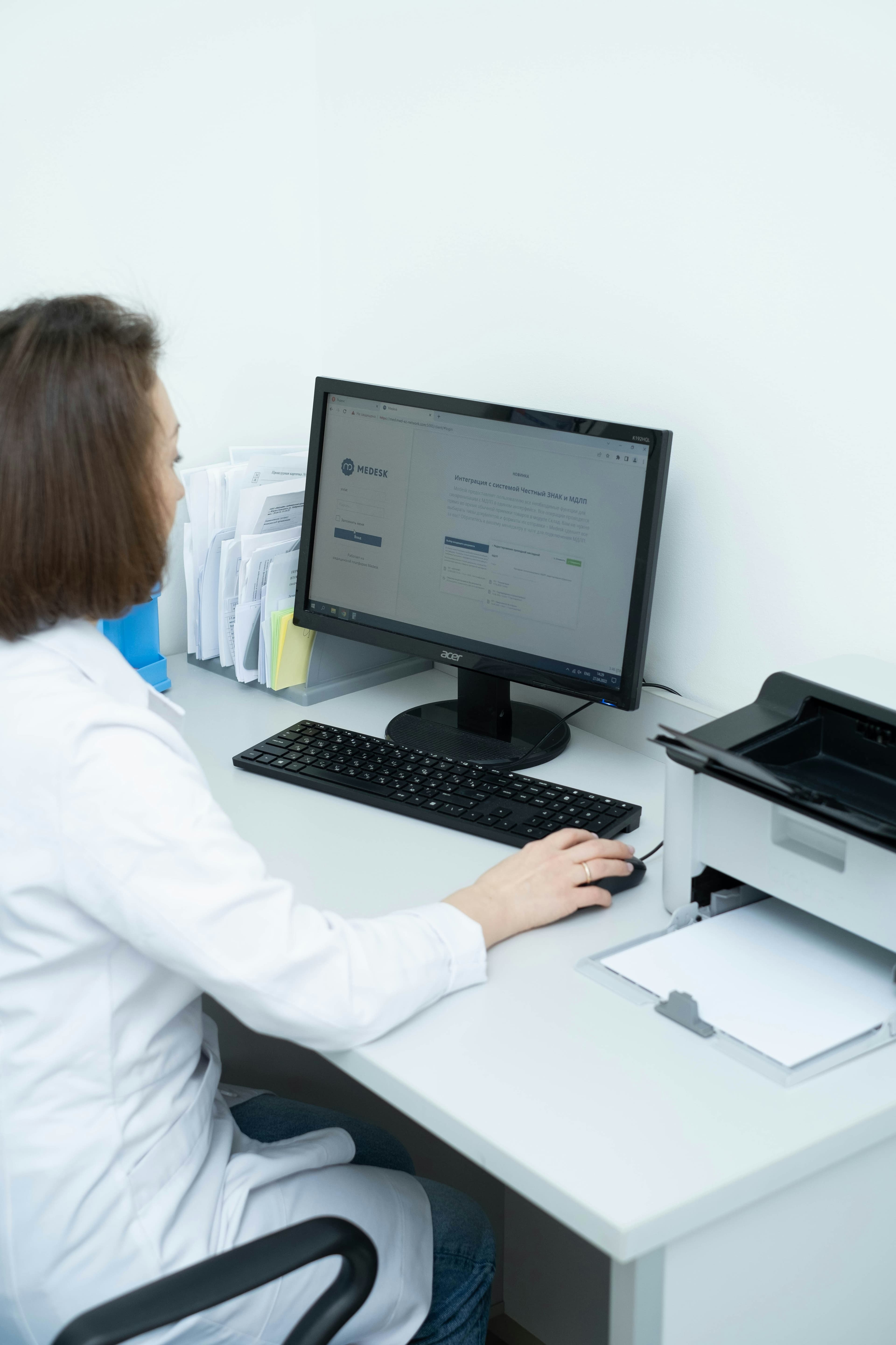 A clinic staff member at her computer with document trays and a printer beside her